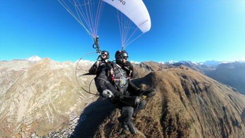 The brothers tandem in the air with a parachute above their heads, with black helmets on, above the French Alps