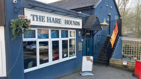 A large blue pub with the Hare and the Hounds sign on the front. There is a hanging basket with pink flowers next to the sign and stairs leading up the outside of the building away from the camera. A white, blank advertising board stands outside.