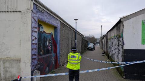 A police officer stood in front of blue and white police tape across the entrance of an alley way with graffiti on the walls.