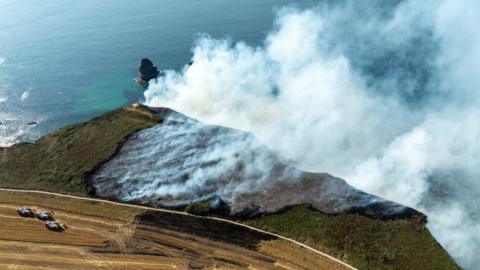 An aerial photo of Basset Cove, Portreath. There is a large amount of smoke on the cliff. The cliff is covered in grass. The sea is in the background.