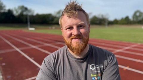 A man with ginger hair and a beard in a grey T-shirt on a running track with grass in the background