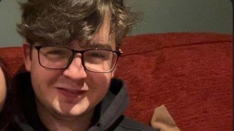 A young man with black rimmed specs and brown wavy hair is smiling. He is in close up on a deep red couch with a grey wall behind.