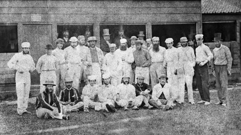 A grainy old black and white photo with 22 men in white outfits, some standing some sitting. All are wearing boater-style hats. Men in top hats can be seen in a spectator stand behind. 