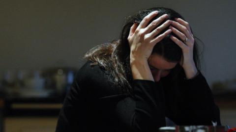 The image shows a woman with her head in her hands. She has dark, long hair and is wearing all black.