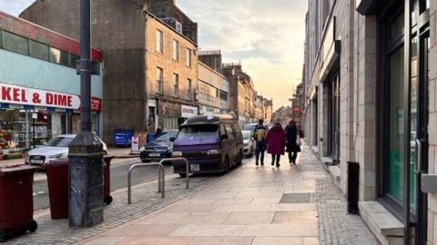Kirkcaldy High Street on a sunny day. There are rows of buildings either side of the street, and a group of people are walking down the pavement.