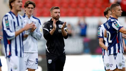 James Morrison, wearing a black West Bromwich Albion tracksuit, stands alongside his players to applaud the fans after the EFL Cup game against Rotherham