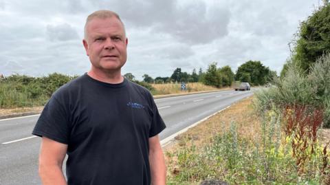 A man wearing a black t-shirt stands in front of a two lane road. The grass and vegetation on the sides of the road are dried out because there has been so little rain. There is a car pulling a trailer in the background.