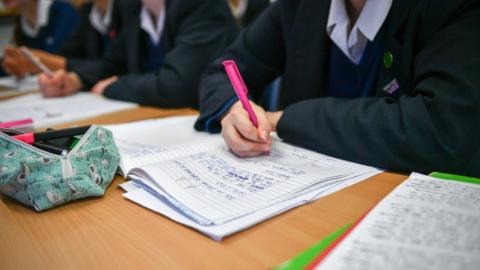 Female pupils are sitting at a desk, we can see their shoulders and arms. They are wearing white shirts, navy V-neck jumpers and navy blazers. They have exercise books open and are writing. A pencil case is in front of the nearest pupil. 