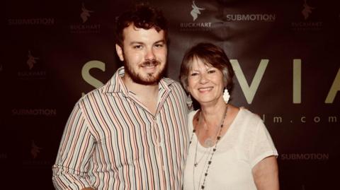 Jonny Hamer wearing a beard and a striped shirt stood in front of a large poster which says Sylvia alongside his mother Heather