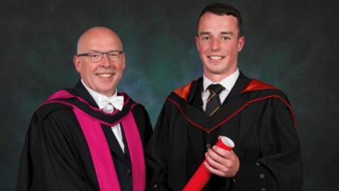 Father and son John and Cameron Gibson, pictured inside at Cameron's graduation. Both are wearing gowns and Cameron is holding his degree.