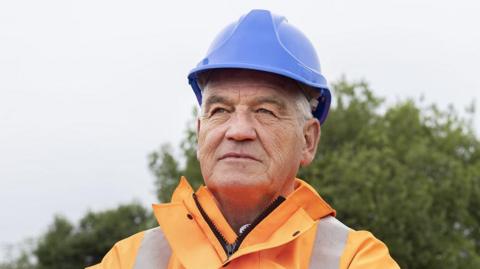 Gateshead Council leader Martin Gannon at the Leamside Line, wearing a blue hard hat and orange hi-vis jacket. He is staring into the distance.