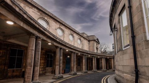 A curved road outside a sandstone building with a columned walkway and round windows above it