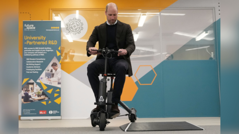Prince William sits on a grey, light weight scooter with three wheels. He is wearing a black suit, black sweater and white shirt. The wall behind is decorated with geometric shapes and cogs, and a banner reads "University-partnered R&D".