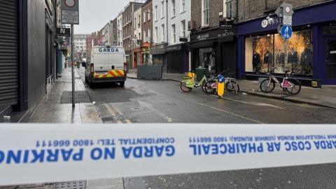 A street is sealed off with Garda tape in Dublin city centre. A Garda van is in the street as are a number of shops and bikes.