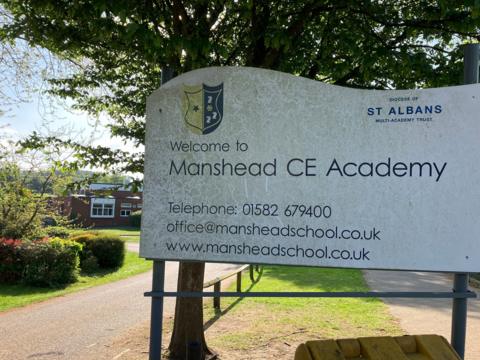 A discoloured white sign for Manshead CE Academy. There is a green tree behind the sign and a driveway to the left of the sign which has details of the school on it.