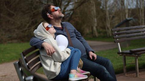 A man and a child watching a partial solar eclipse at Keele university on 29 March, 2025. They are wearing special glasses and are sat on a bench and there is part of what looks like an empty bench next to them. There is grass in the background. 