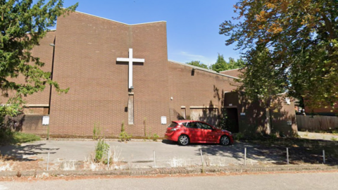 St Thomas' Church is a brown bricked building which has a silver cross outside the front. On the right is a doorway with a small red car out the front.
