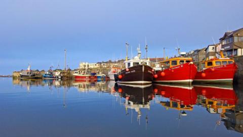 Fishing boats moored at the harbour at Seahouses. Their reflection can be seen in the water. There are three berthed in the foreground with more behind. Set back from the harbour wall on the right is a long row of houses and other buildings which face out to sea.