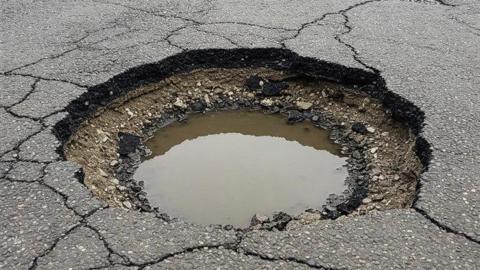 General view of a pothole filled part-way with water surrounded by cracked tarmac