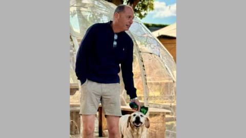 Robin Cooper looking to the right and petting a labrador with one hand. He is smiling and wearing casual clothes. 