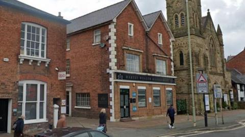 Two red brick buildings to the left of a stone church, with people walking on the street next to the buildings.
