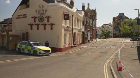 A police car parked outside a pub in Newton Abbot. The street has been cordoned off. A police officer is stood next to the police car.