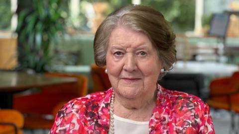 Patricia Routledge sits indoors wearing a pink, red and white patterned jacket and a pearl necklace. The photograph was taken in a bright room with large windows and orange chairs visible in the background.