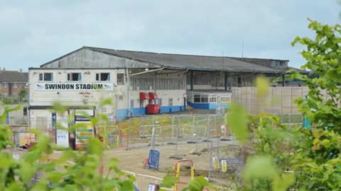 The edge of Swindon Abbey Stadium. There is a white and blue building with a metallic roof. At the side of the building hangs a white banner with black writing reading 'Swindon Stadium'. There are silver fences joined together in front of the stadium.