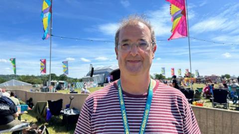 Graeme Merifield, a middle-aged man is stood on the festival site wearing a stripey t-shirt and glasses. 
