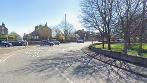 A road scene taken on a clear, sunny day. The viewpoint is from street level, looking toward a curved section of the roundabout. Several cars are travelling in both directions.
