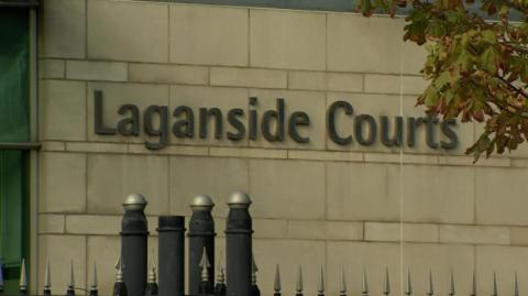 The exterior of a white stone exterior building with a black iron gate in the foreground. There is a sign on the side of the stone building that says 'Laganside Courts' in silver letters.