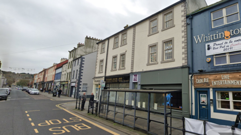 A high street which has Georgian terraced houses on it. There is a pub and other businesses on the bottom row of the houses. There is a large bus stop and a bus shelter with two people waiting under it.