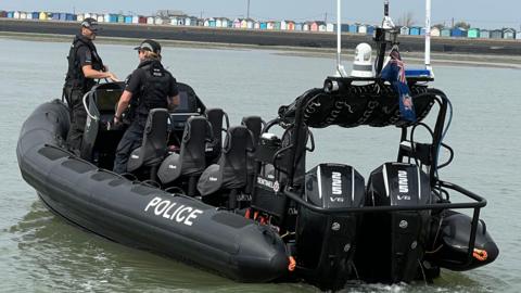 A wide shot of a black boat out on the water with the word police written along the side. Two officers are on board