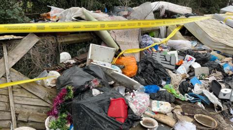A pile of rubbish including mattresses, black bin bags is covered in yellow tape.