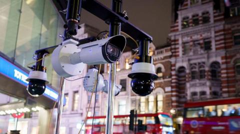 Close-up of security cameras mounted on a metal rig outside a London Underground station at night, with red double-decker buses and pedestrians in the blurred background.
