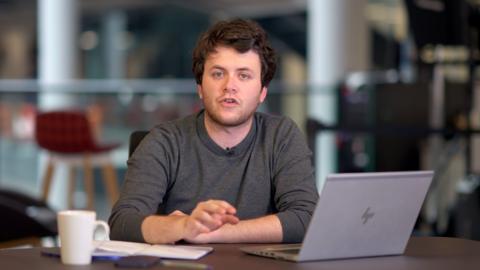 BBC journalist Ben Summer, a young man with brown hair and stubble speaking to the camera while sitting with a laptop and mug at a desk