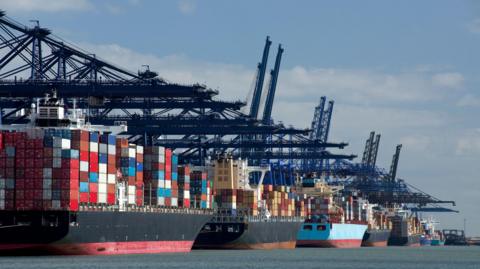 Six boats carrying containers are lined up in the sea. Above them are blue cranes.