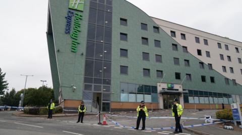 A large green and white building with a vertical "Holiday Inn Express" sign mounted on the side. Four police officers stand outside the hotel next to blue and white police tape. Behind the tape, bricks and debris is lying in the ground.