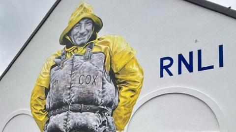 A mural in yellow and grey of Thomas James King in waterproof clothing on a white wall with a large blue RNLI sign next to it.