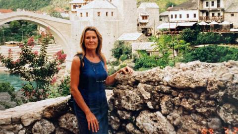 Ann is wearing a blue top and blue denim skirt and standing in front of the reconstructed Stari Most, also known as the Old Bridge in Mostar, Bosnia.