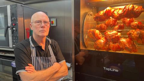 Steve Moloney stands with his arms crossed wearing a striped apron while stood next to an oven filled with rotisserie chicken. 