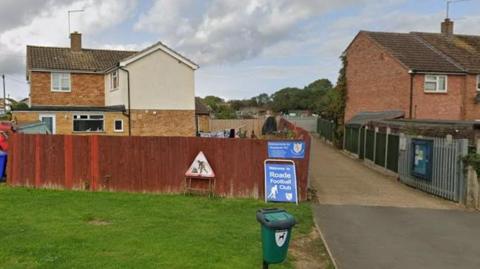 Entrance off residential street into football ground, with blue Roade FC sign