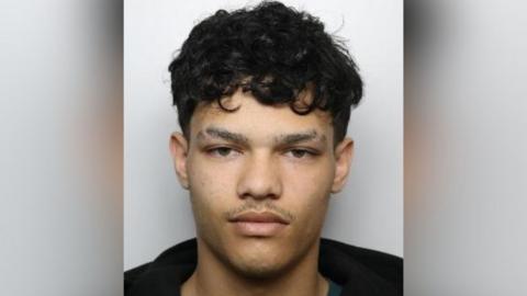A mugshot of a man with short curly black hair and slits cut out of his eyebrows. He has a thin moustache and wears a black hoodie.