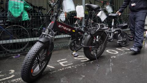 A modified e-bike, seized by police, is propped up by a kick-stand. There is a man behind wheeling a smaller e-bike and to the side opposite the camera there is a storage area containing a number of other bikes.