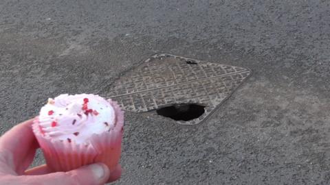 A man holding a pink cupcake in front of a manhole cover with a very large hole in it.