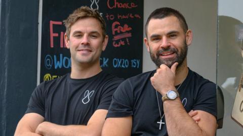 The Talbot brothers standing outside one of their salons. They wear black T-shirts, possibly the salon uniform. George has short brown hair. James' hair is more black, and he has a dark beard. He wears a cross around his neck.