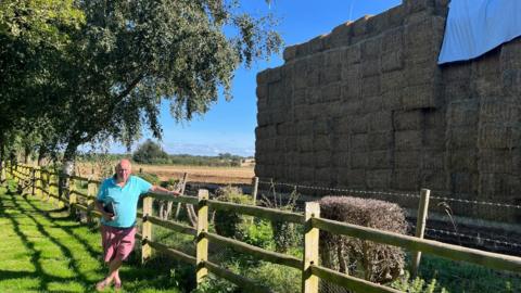 Cliff Nye is wearing a blue polo shirt and pink shorts and standing next to a boundary fence on his property, dwarfed by a giant straw structure in the field next door.