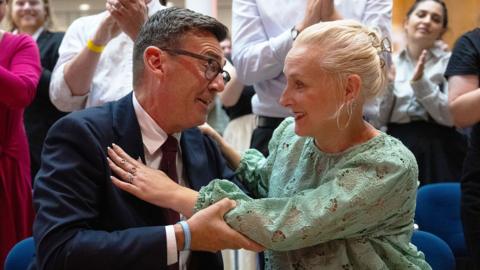 Andy Burnham hugging his wife, Marie-France van Heel. Burnham is wearing a dark suit with a shirt and tie and glasses. Van Heel is wearing a green dress. She has blond hair and hooped earrings. 