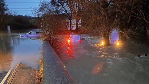 Three vehicles - two vans and a Range Rover - are half submerged in floodwater in Axminster, Devon, during Storm Chandra. The two vans have their lights on. The photo was taken during the early hours of the morning. Several trees and a house are in the background.
