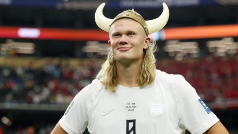 Erling Haaland, Norway's star striker, stands with his hands on his hips looking out towards the crowd of a football stadium, smirking. He's wearing a white jersey with the number 9 printed on its front. A plastic viking helmet with two white horns on each side rests atop his head.